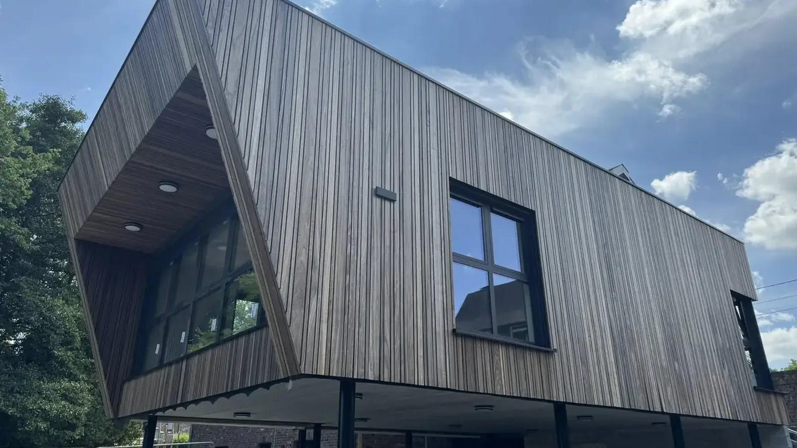 Bâtiment en bardage de bois à lames verticales, posé en encorbellement sur des poteaux, sous un ciel nuageux