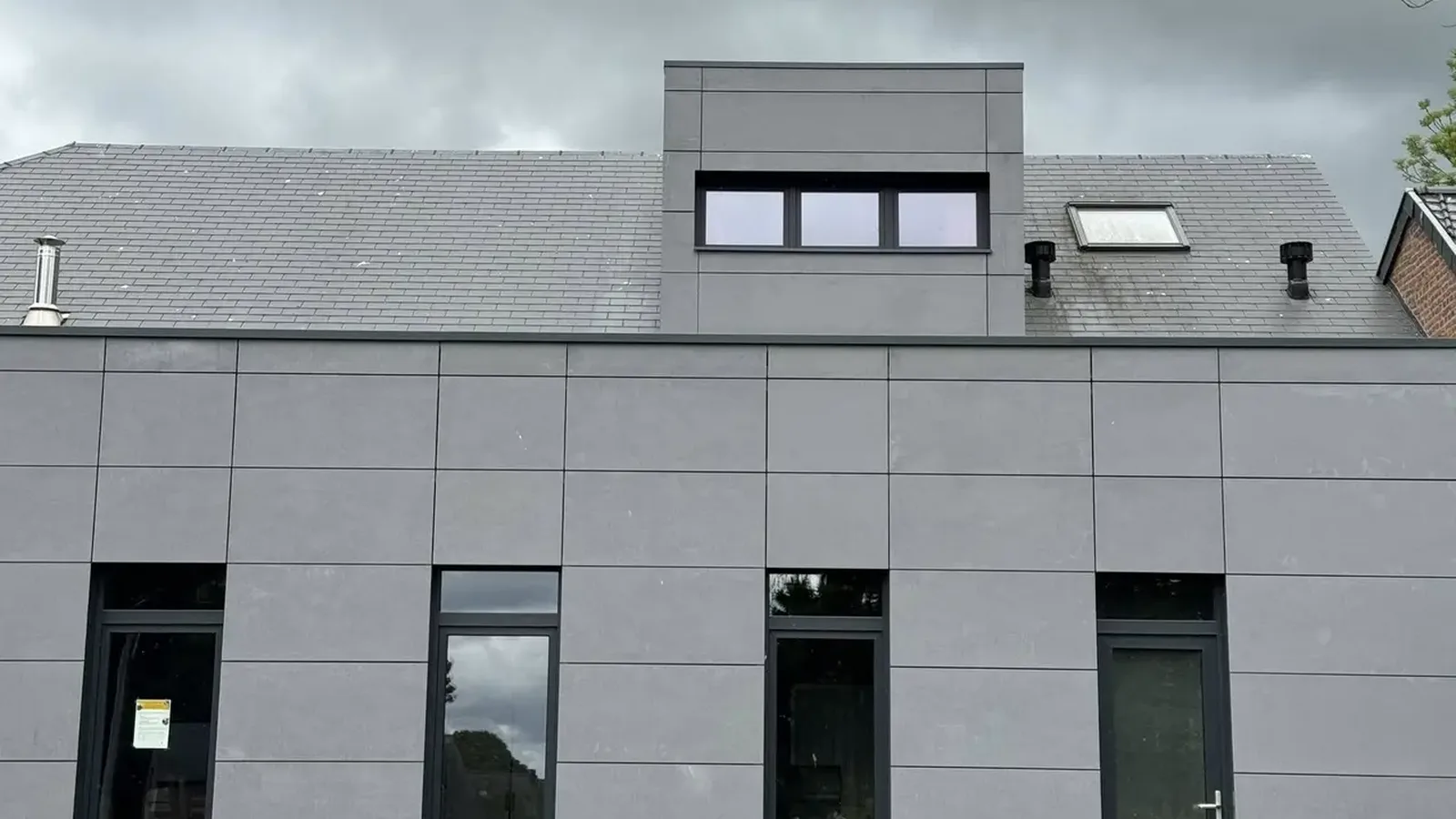 Façade en grands panneaux gris clair avec fenêtres sombres sous un toit d'ardoises