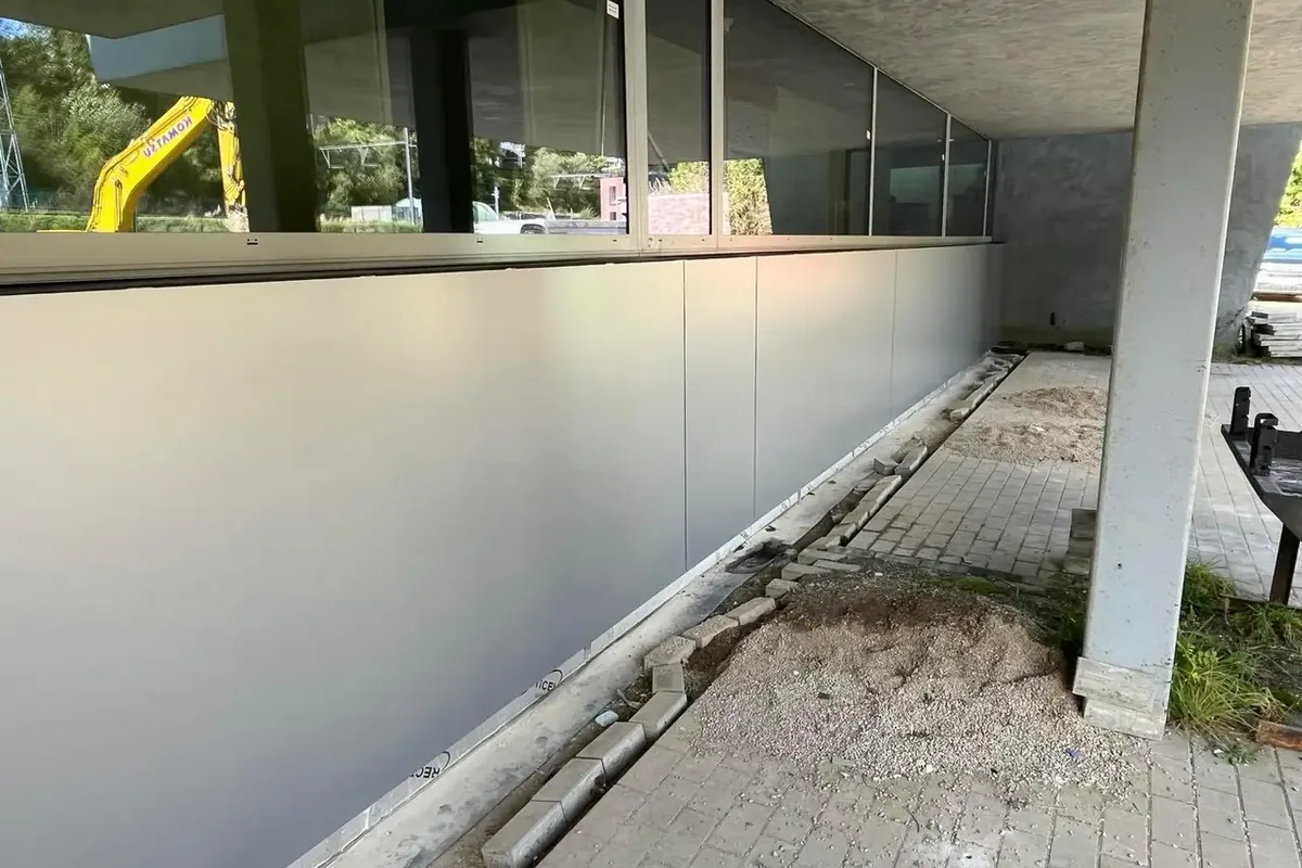 Long mur de panneaux lisses gris clair sous un auvent, côté chantier