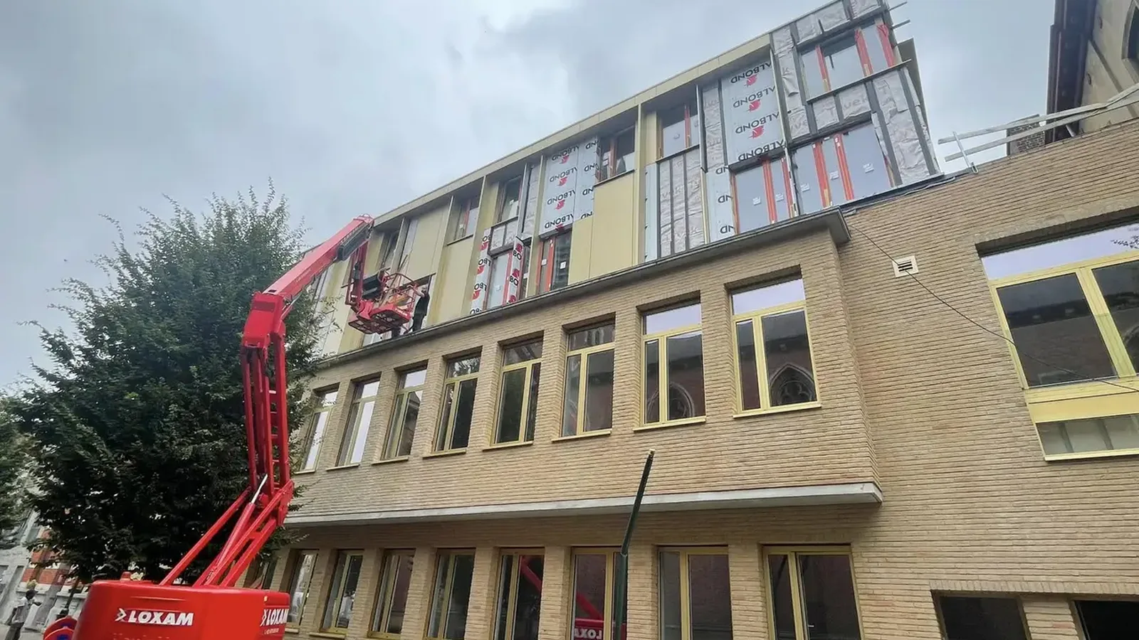 Immeuble ancien en briques dont la partie haute reçoit des panneaux isolants et un habillage, avec une nacelle rouge devant