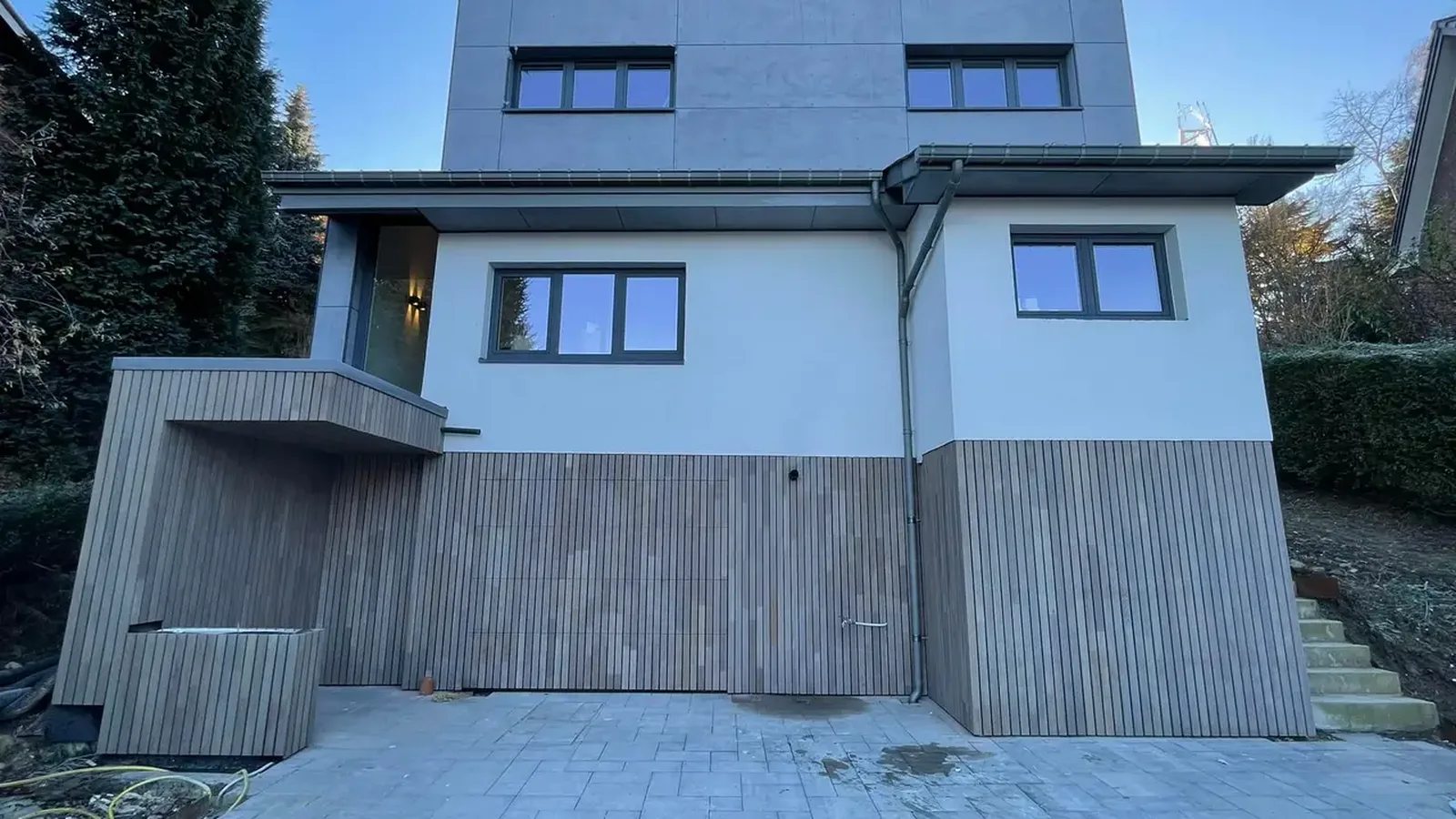 Maison à façade enduite blanche, avec soubassement en lames de bois grisées, sous un ciel bleu