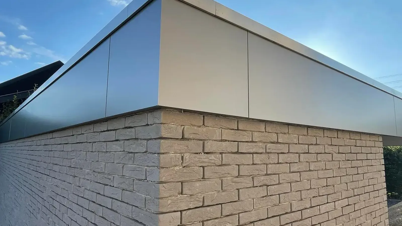 Angle d'une annexe en briques claires coiffée d'un bandeau d'aluminium gris, sous un ciel bleu